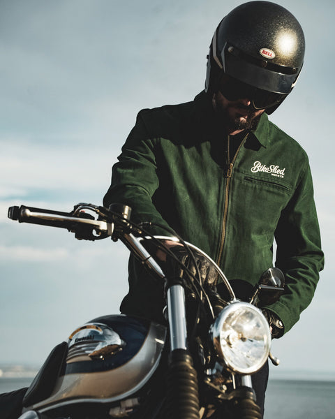 Model wearing the Bike Shed Moto Co. Twill Green logo jacket standing next to a motorcycle.
