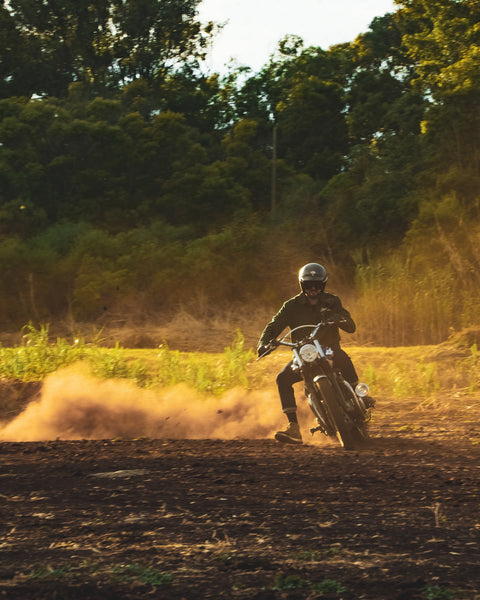 Model wearing the Bike Shed Moto Co. Twill Green logo jacket a motorcycle near the woods.