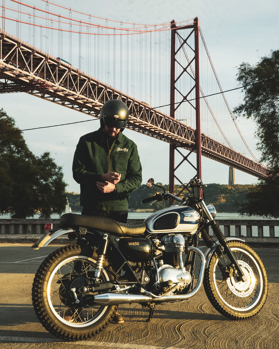 Model wearing the Bike Shed Moto Co. Twill Green logo jacket standing next to a motorcycle.