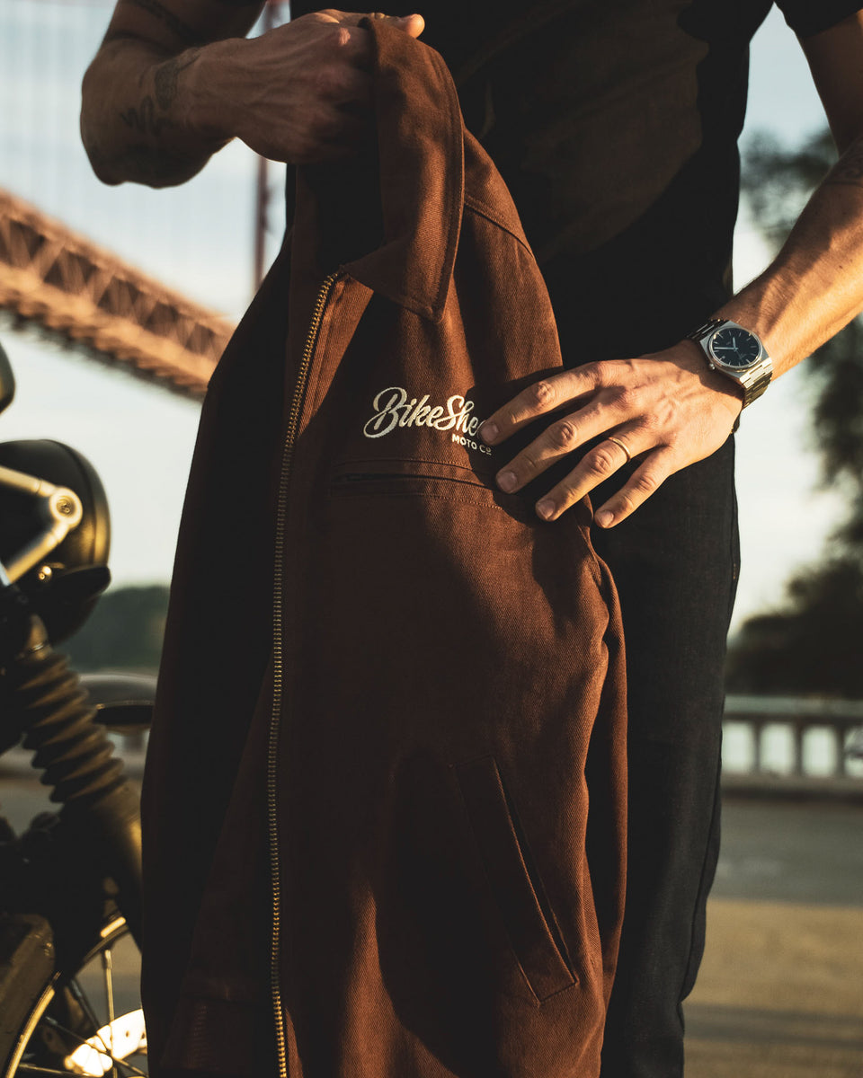 Model holding the Bike Shed Moto Co Twill brown jacket with a Bike Shed brand logo.