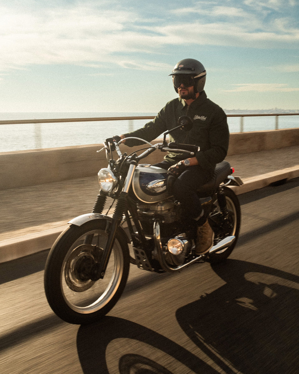 Model wearing the Bike Shed Moto Co. Twill Green logo jacket riding a motorcycle.