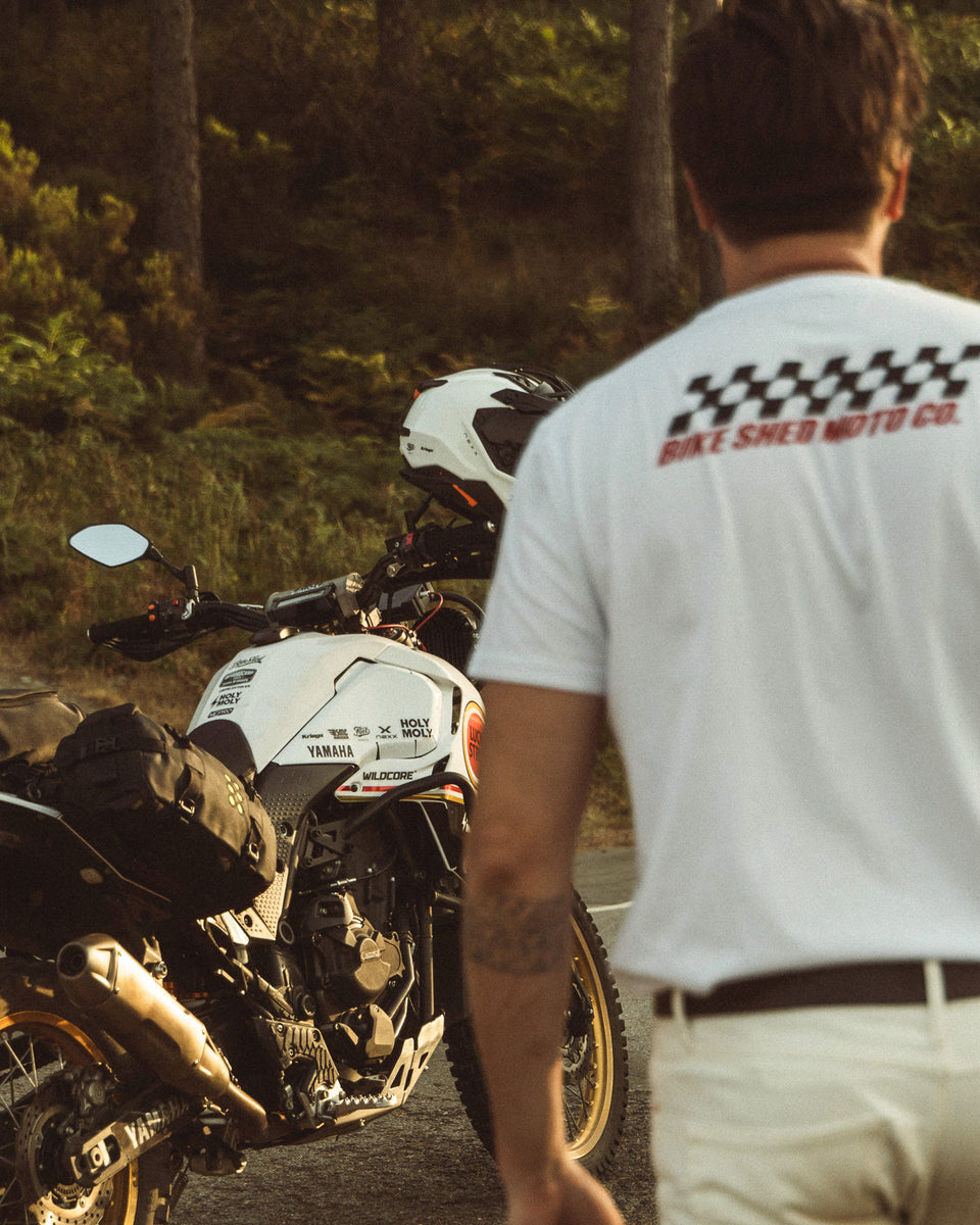 Model wearing Motorcycle Desert Race white t-shirt with a Bike Shed Moto logo, standing next to  a motorcycle in a forest.