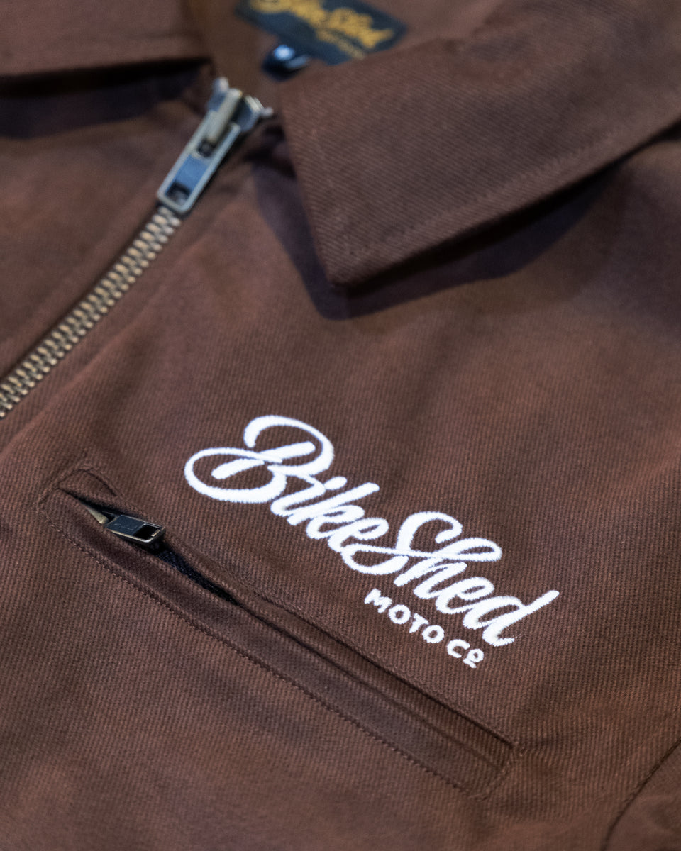 Brown twill motorcycle jacket with 'Bike Shed Moto Co' logo on a close-up of the chest area.